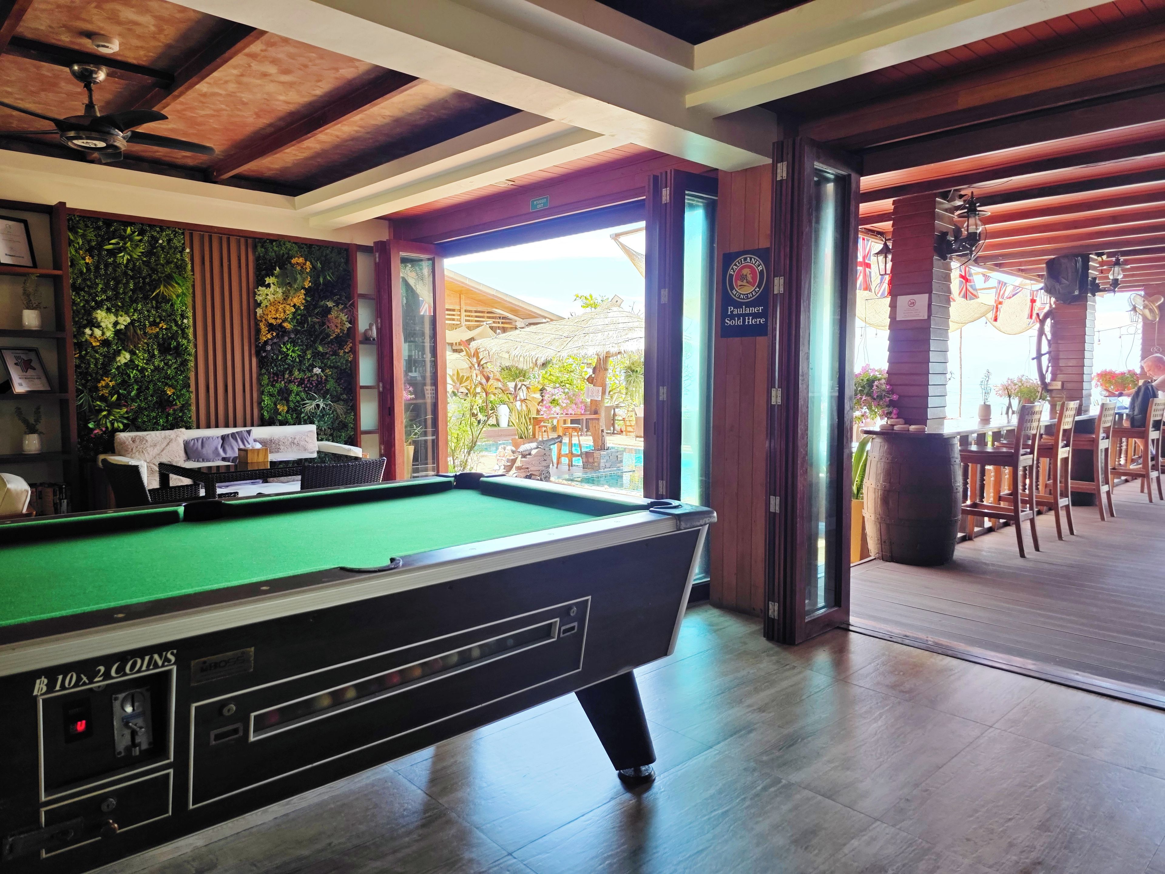 A pool table in a room with wooden beams, a sofa, and a view of an outdoor area with a pool and umbrellas.