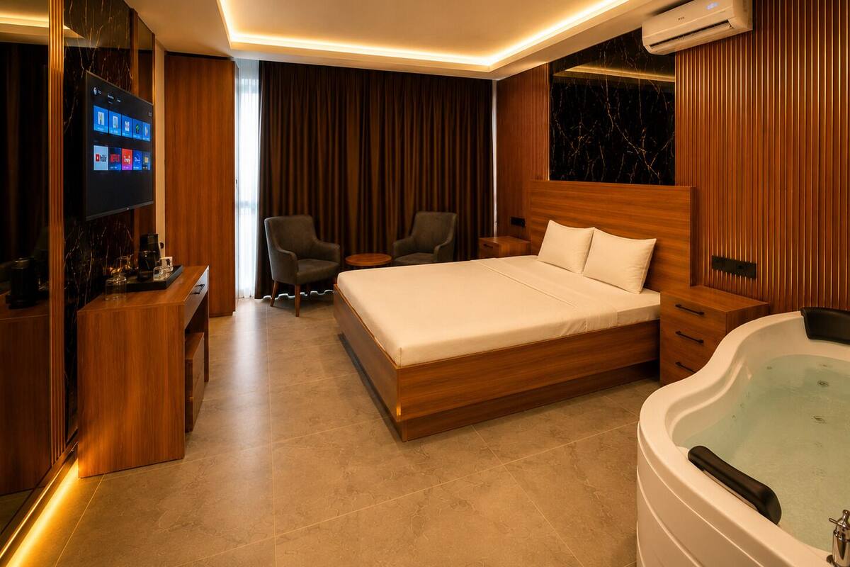 A modern hotel room with a large bed, a sitting area with chairs, a TV, a minibar, and a jacuzzi.