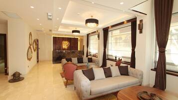 A spacious living area with a sofa, a coffee table, and a dining table set for four. A reception desk with a marble countertop and a logo is visible in the background.