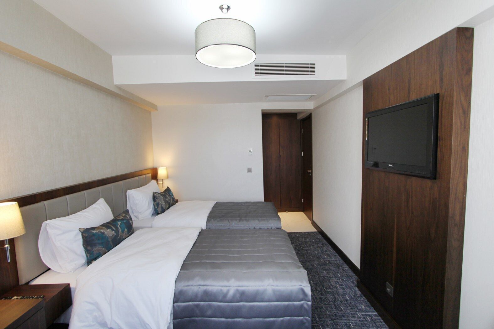 A hotel room with two beds, a flat-screen TV, a ceiling light, and a wooden wardrobe.