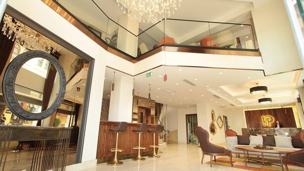 Un vestíbulo de hotel con techo alto, un candelabro, una mesa de recepción y un área de descanso con sofá y sillones.