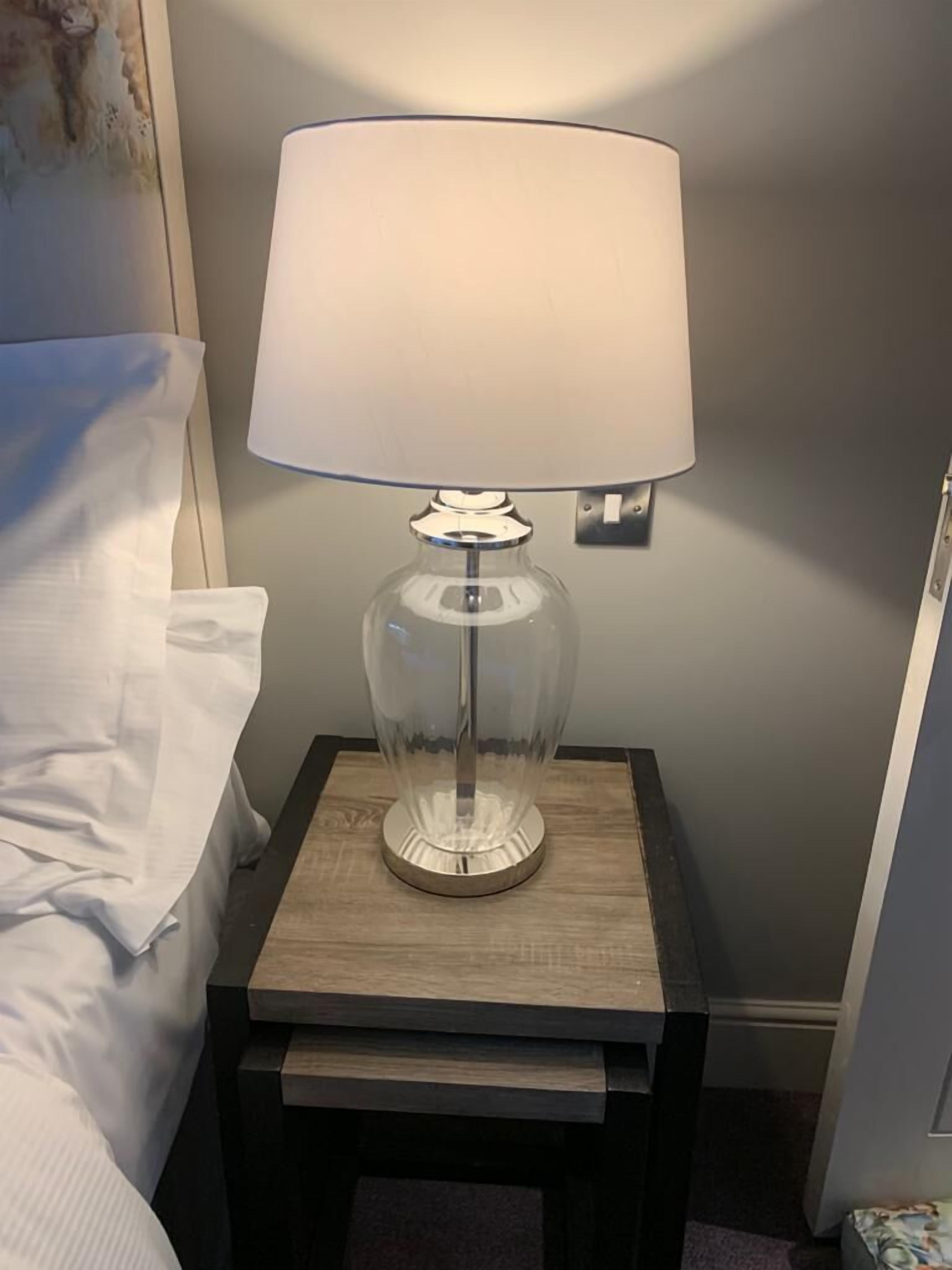 Una cama con ropa blanca, una mesita de noche con lámpara y una puerta a la derecha.