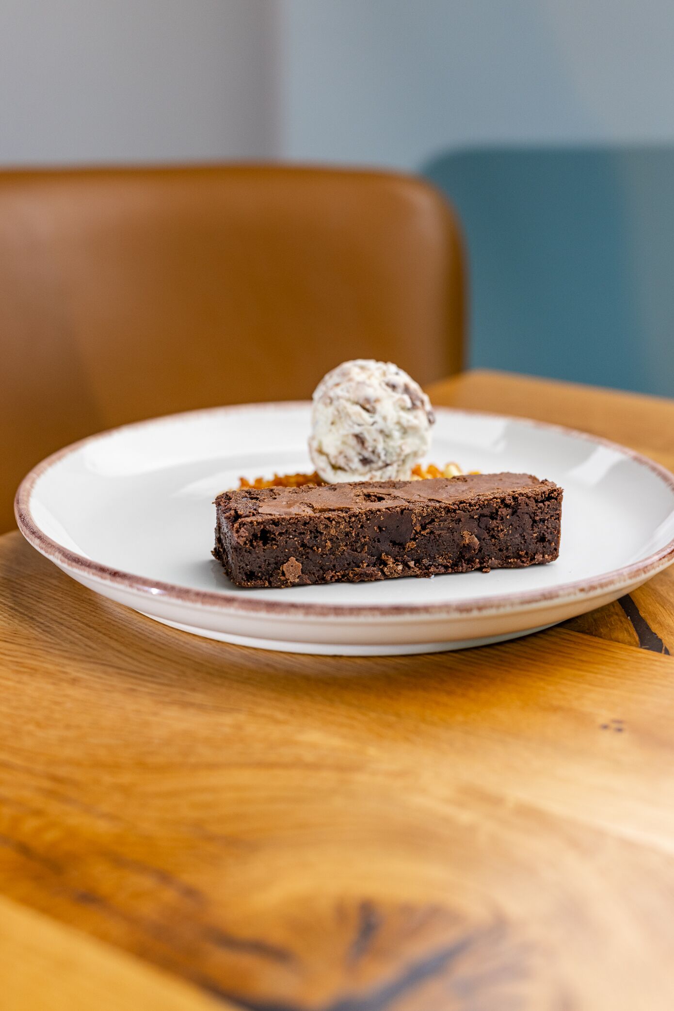A dessert plate with a brownie and a scoop of ice cream on a white plate with a brown rim, placed on a wooden table.