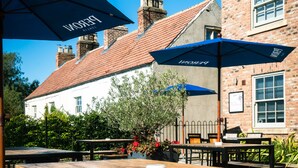 An outdoor seating area with wooden tables and blue umbrellas, set against a brick building with a red-tiled roof. - Plum and Partridge Inn (York)