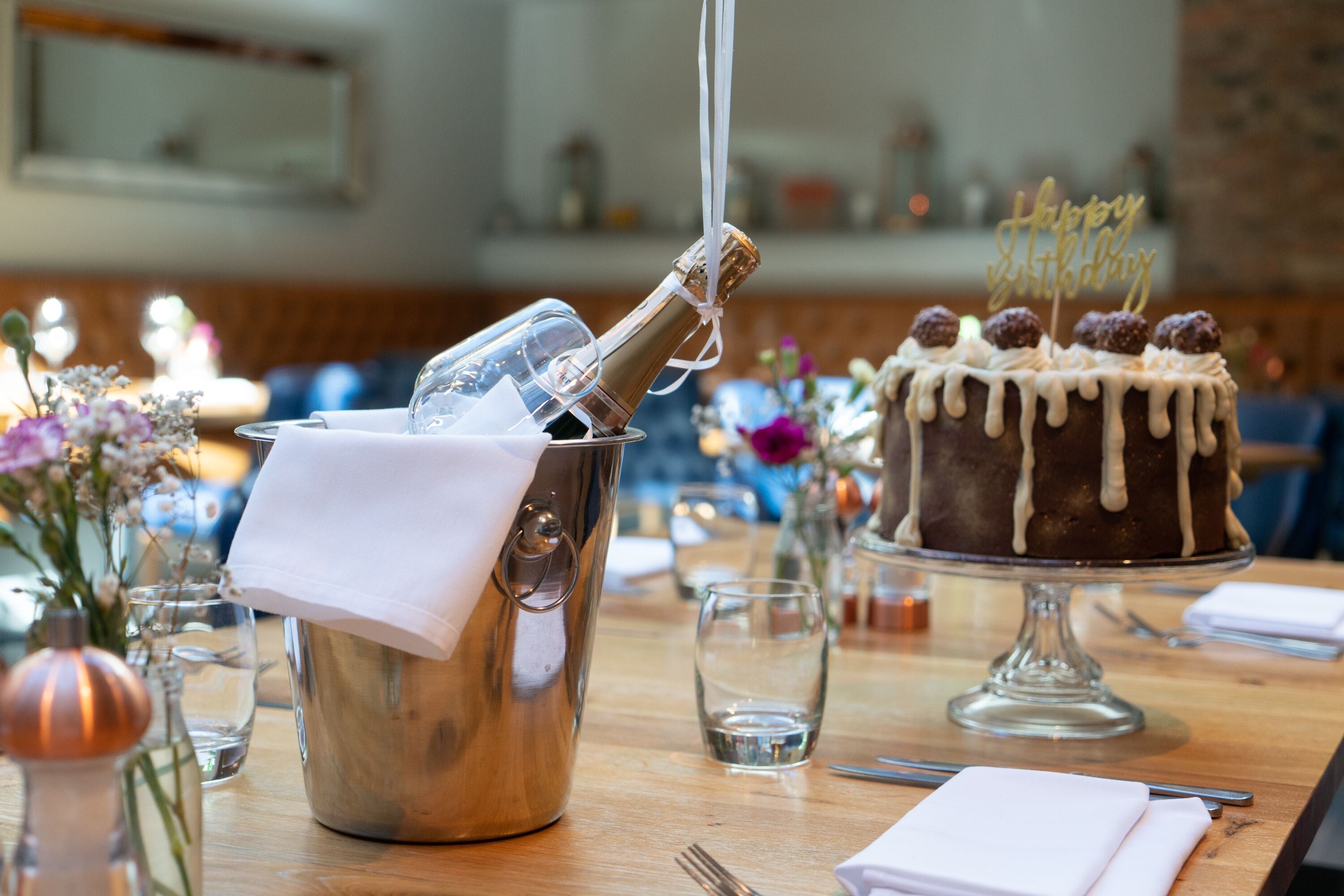 Una mesa de comedor con una botella de champán, un pastel con la inscripción 'Happy Birthday' y un jarrón de flores.