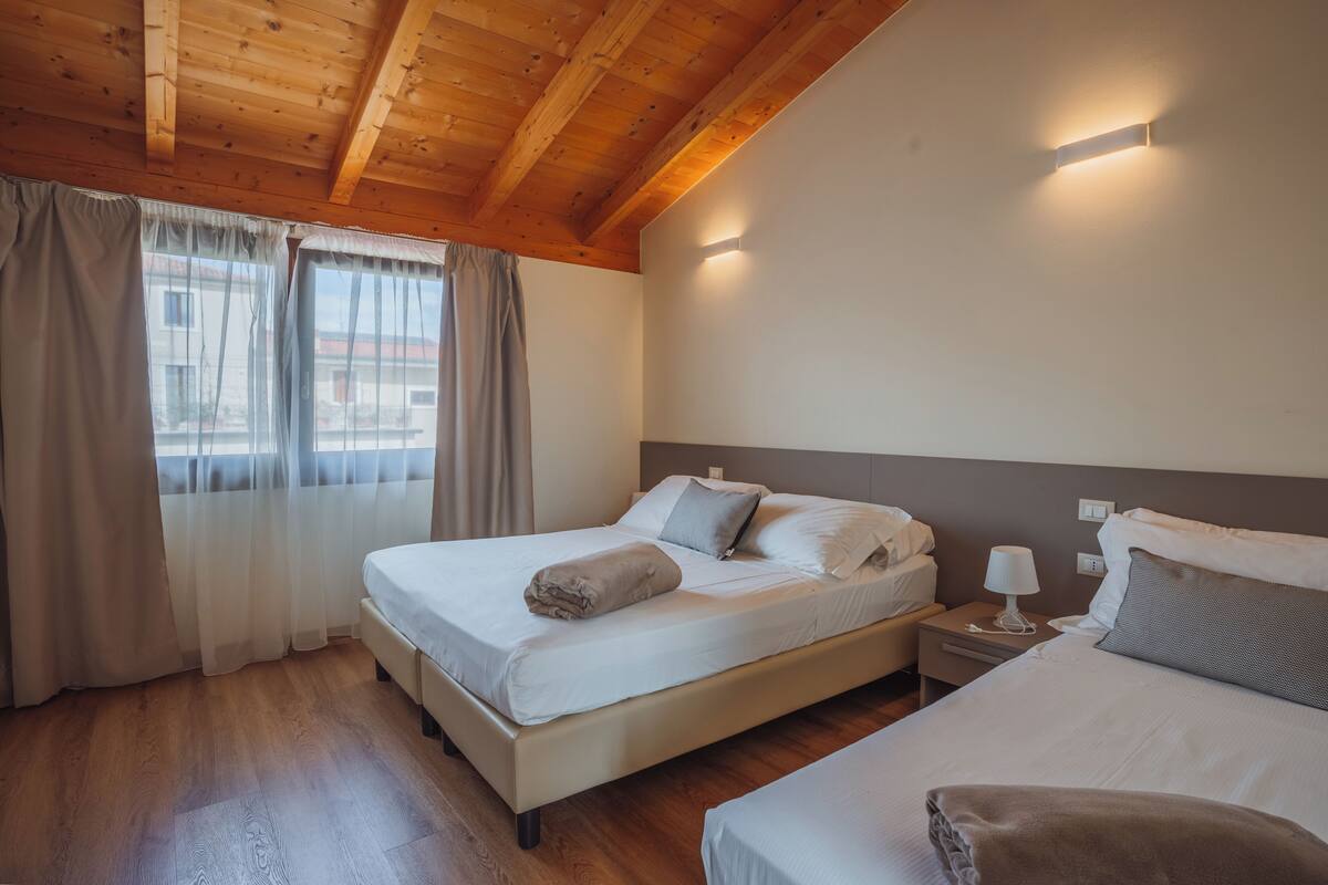 A bedroom with a wooden ceiling, a bed with white bedding, a nightstand with a lamp, and a window with curtains.