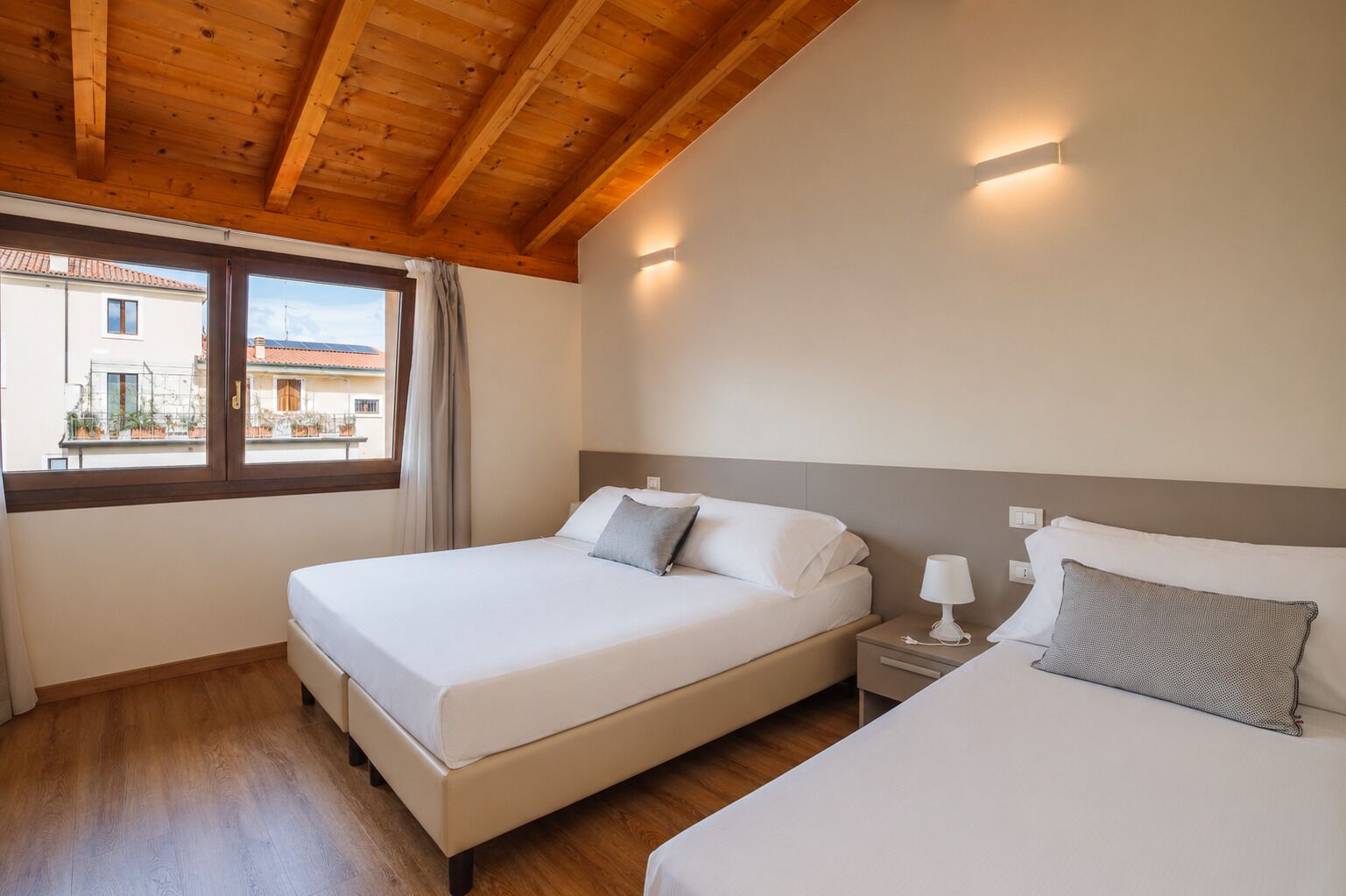 A hotel room with two beds, wooden flooring, a window with curtains, and a view of buildings outside.
