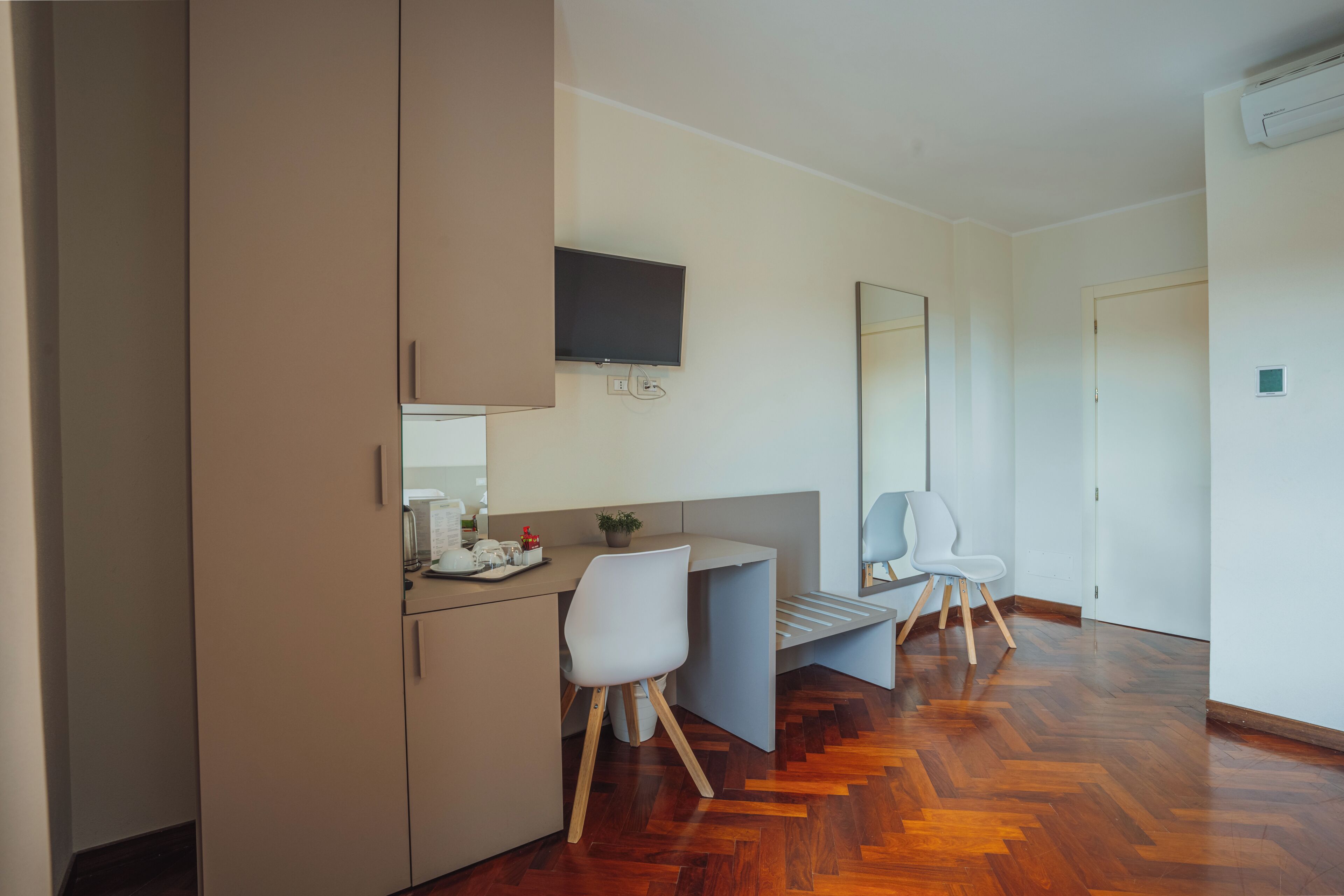 A modern hotel room with a wooden herringbone floor, a kitchenette with a flat-screen TV, a large mirror, and a seating area with a white chair.