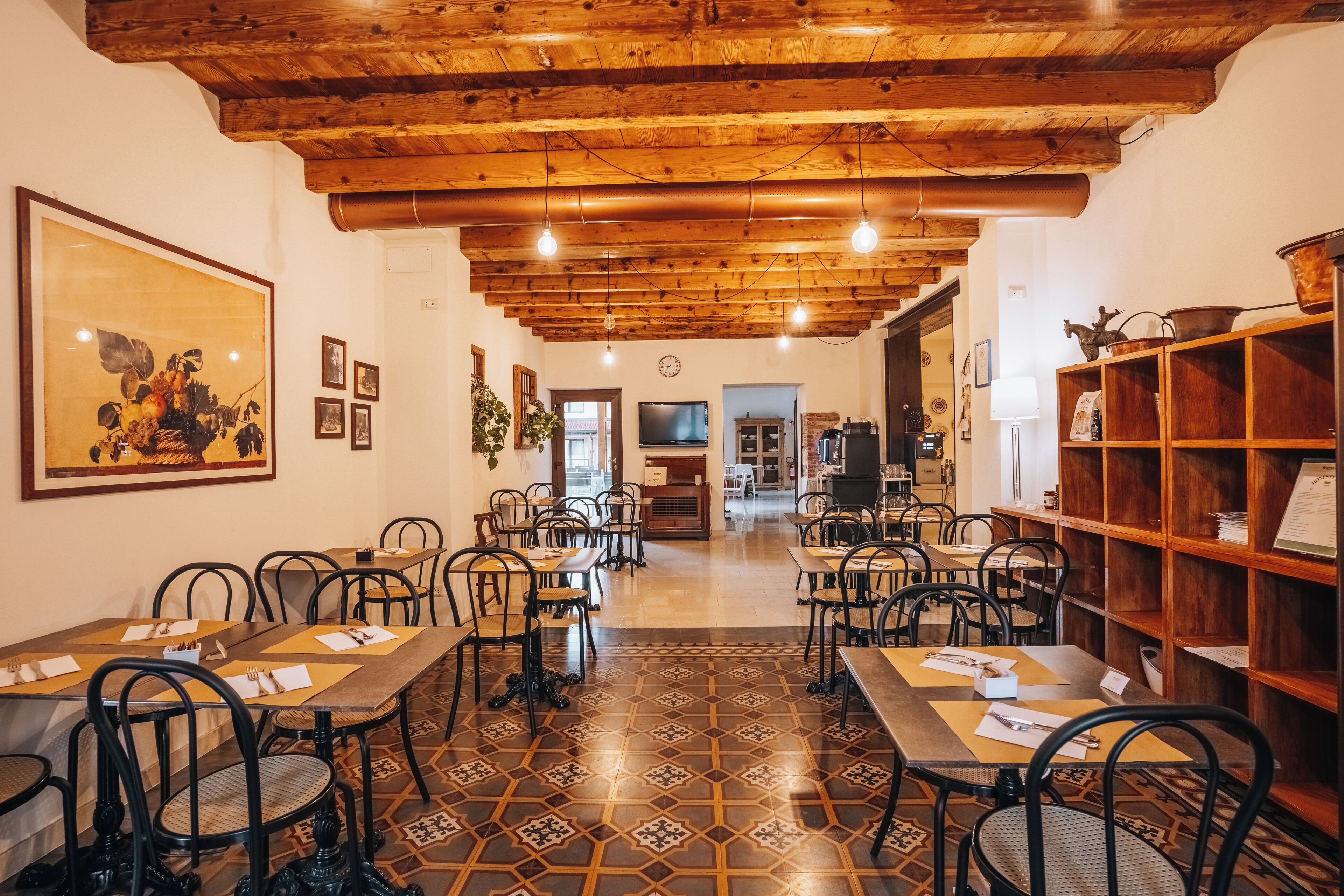Ein Restaurant mit Holzbalkendecke, gemusterten Bodenfliesen und Metallstühlen. Die Tische sind mit Untersetzern und Besteck gedeckt.