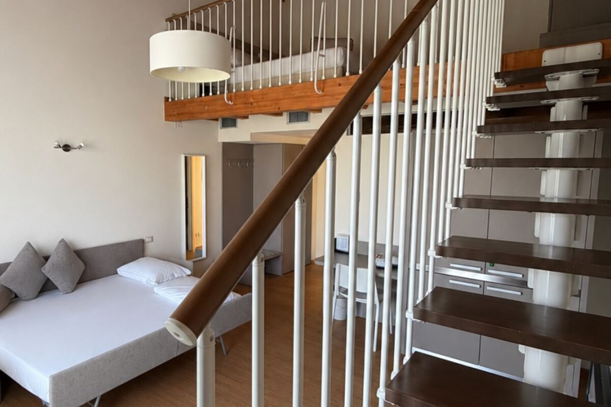 Una habitación moderna con cama, una mesita, una escalera con barandilla de madera y una lámpara colgante.