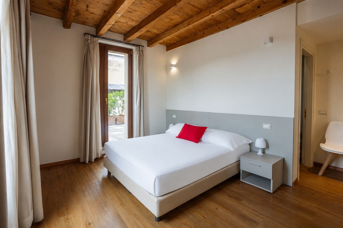 Un dormitorio con una cama grande, una mesita de noche, una lámpara y vistas al exterior.
