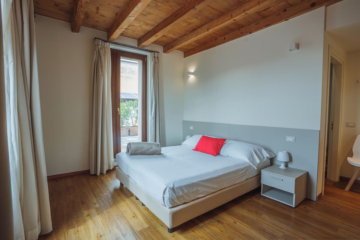 A bedroom with a wooden ceiling, a bed with white bedding and a red pillow, a nightstand with a lamp, and a window with curtains.