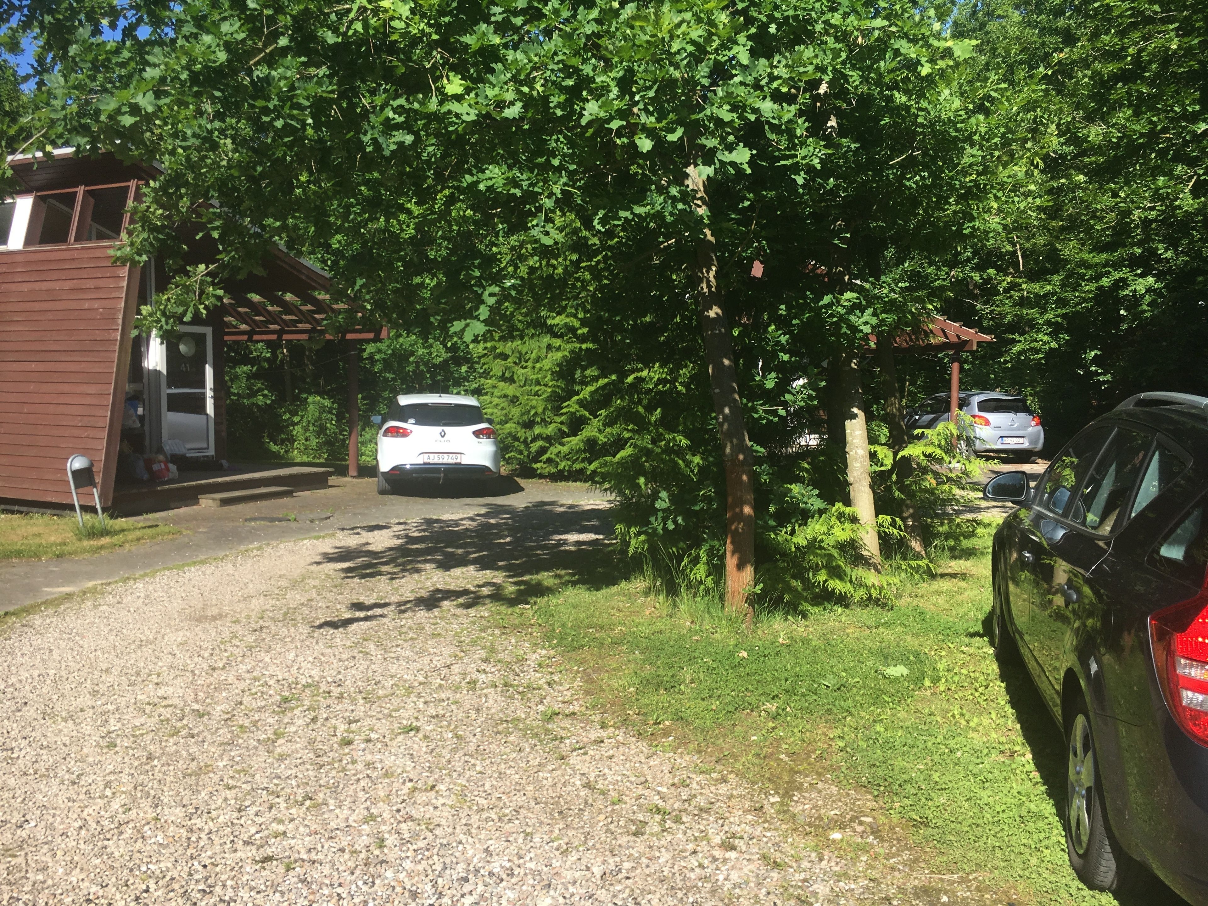 Un camino de grava conduce a un coche blanco estacionado en un área boscosa, con un pequeño edificio a la izquierda.