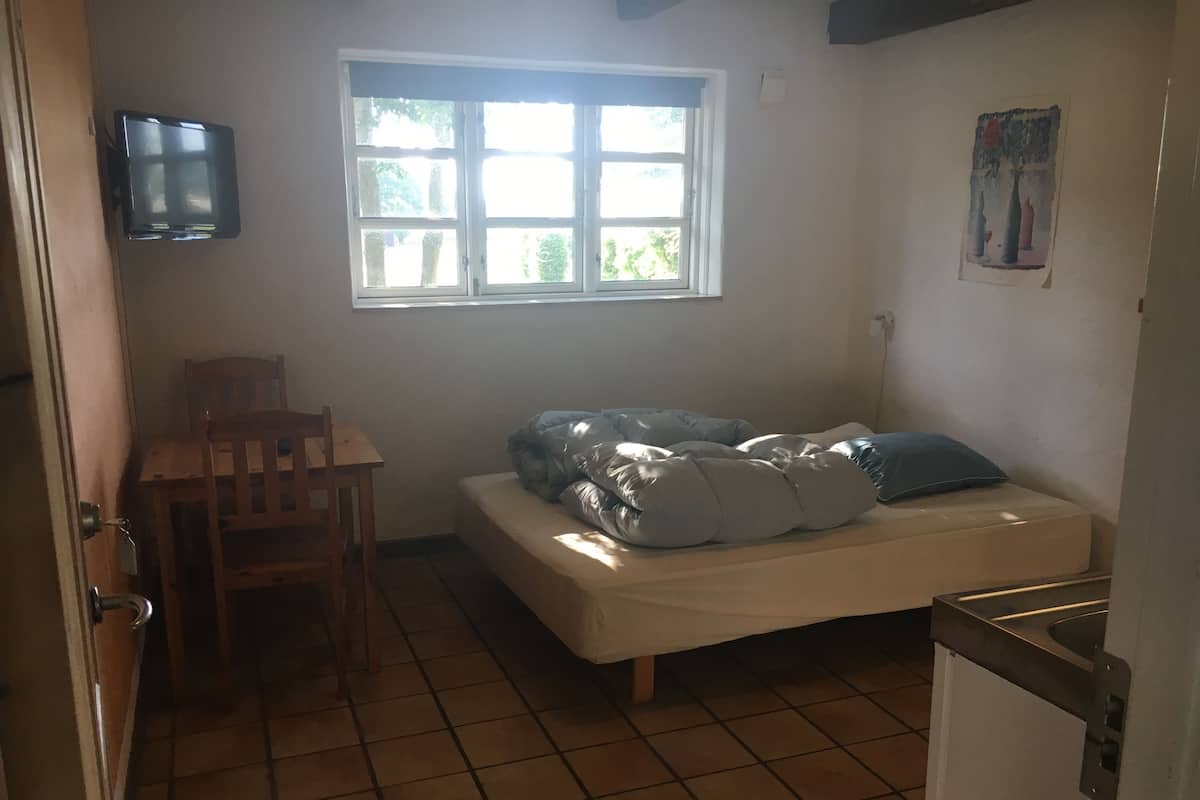 A small, sparsely furnished room with a single bed, a chair, a window, and a wall-mounted TV.