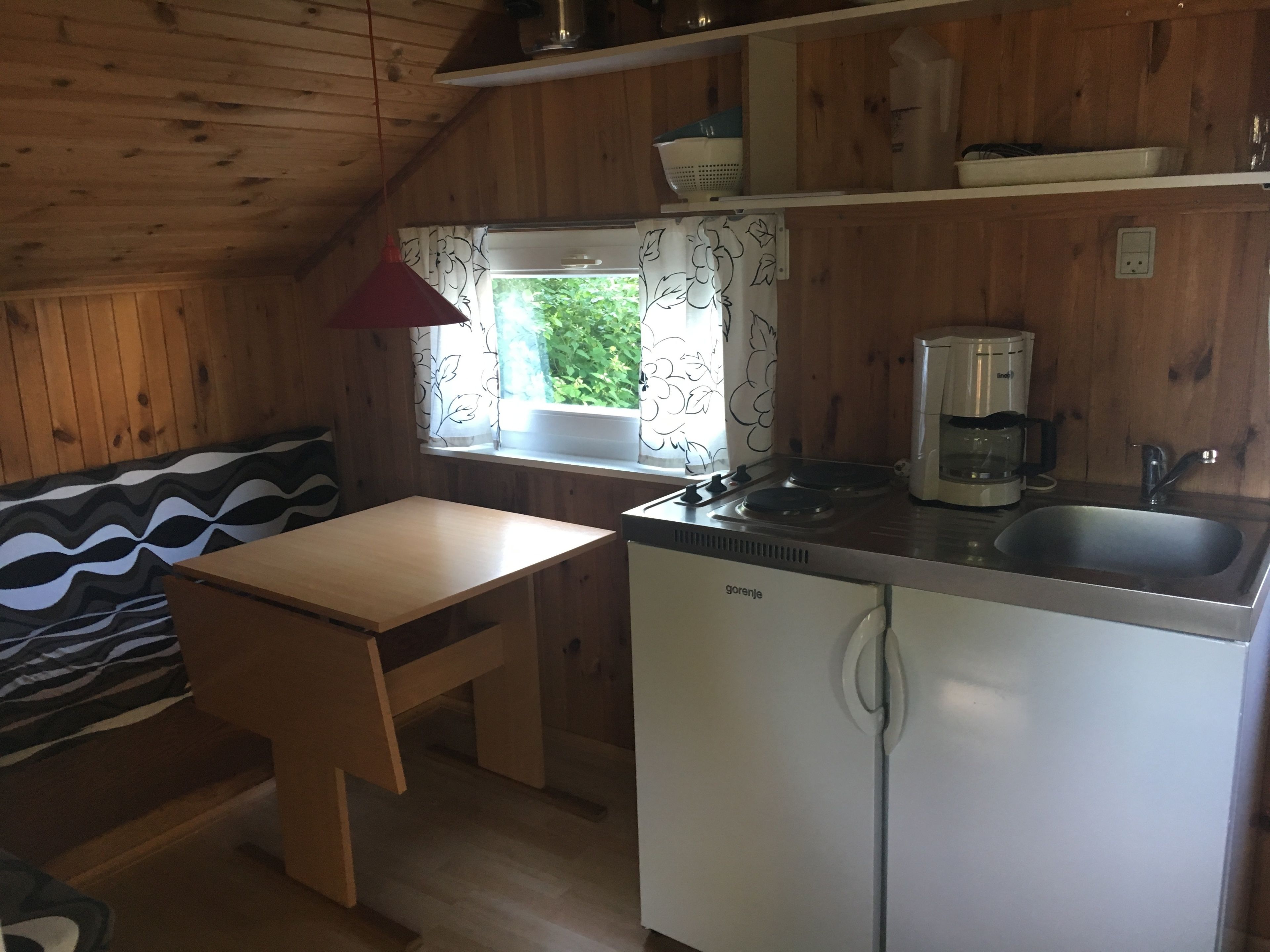 A compact kitchen with wooden walls, a window with curtains, a sink, a coffee maker, and a refrigerator.