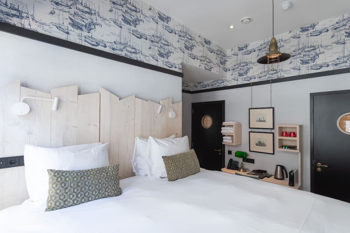 Quarto de hotel moderno com uma cama grande, cabeceira de madeira e papel de parede com tema náutico.