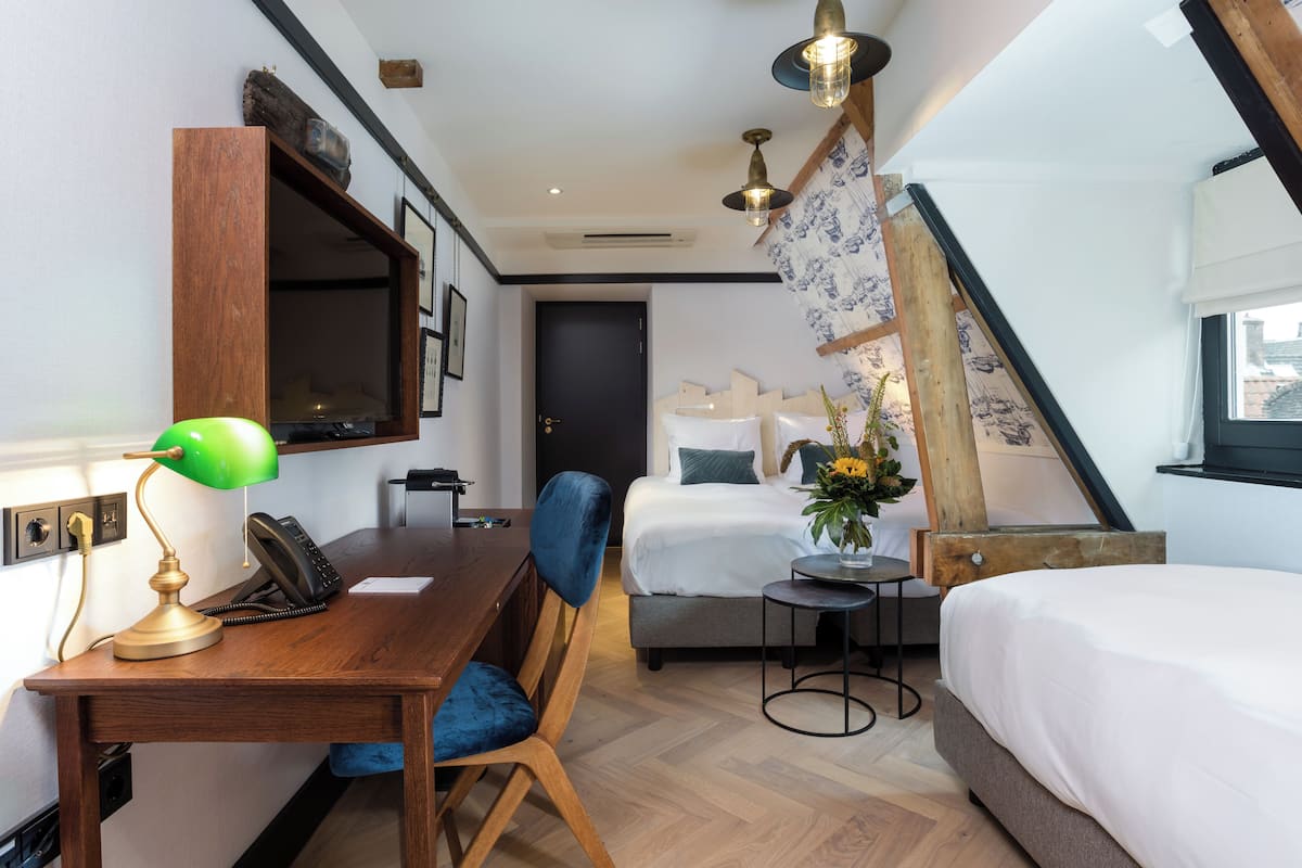 Quarto de hotel compacto com escrivaninha de madeira, uma cadeira, cama, mesa pequena e um vaso decorativo com flores.