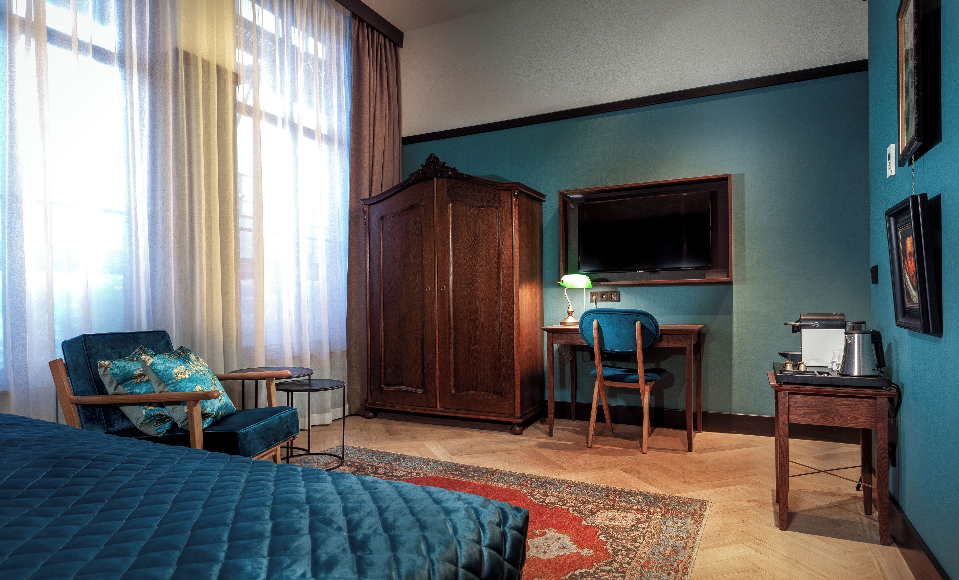 Habitación de hotel con cama, escritorio y silla, armario, televisor y una pequeña mesa con lámpara.
