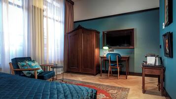 Habitación de hotel con cama, escritorio y silla, armario, televisor y una pequeña mesa con lámpara.