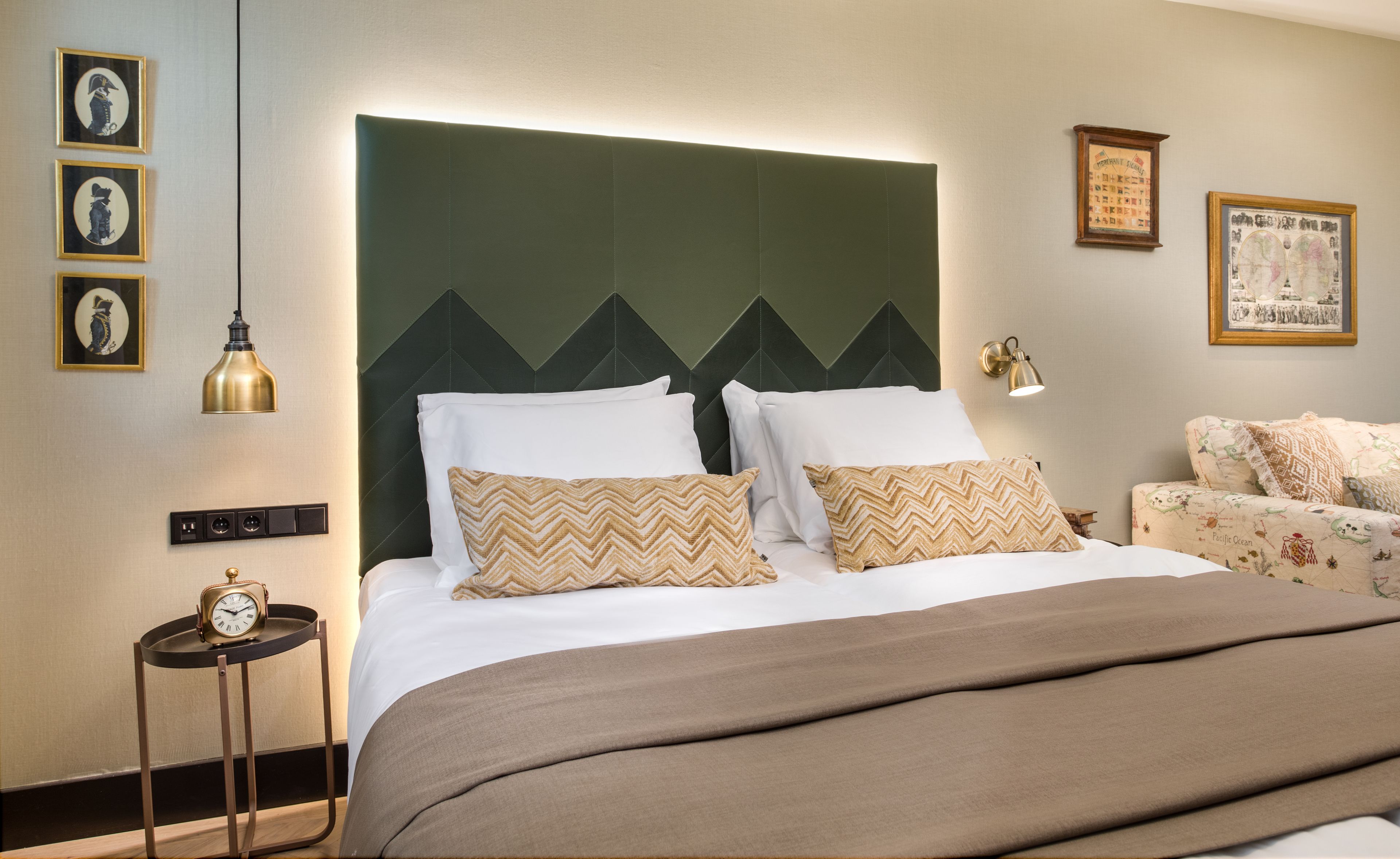 A hotel room with a large bed, a nightstand with a clock, a wall-mounted lamp, and framed artwork.