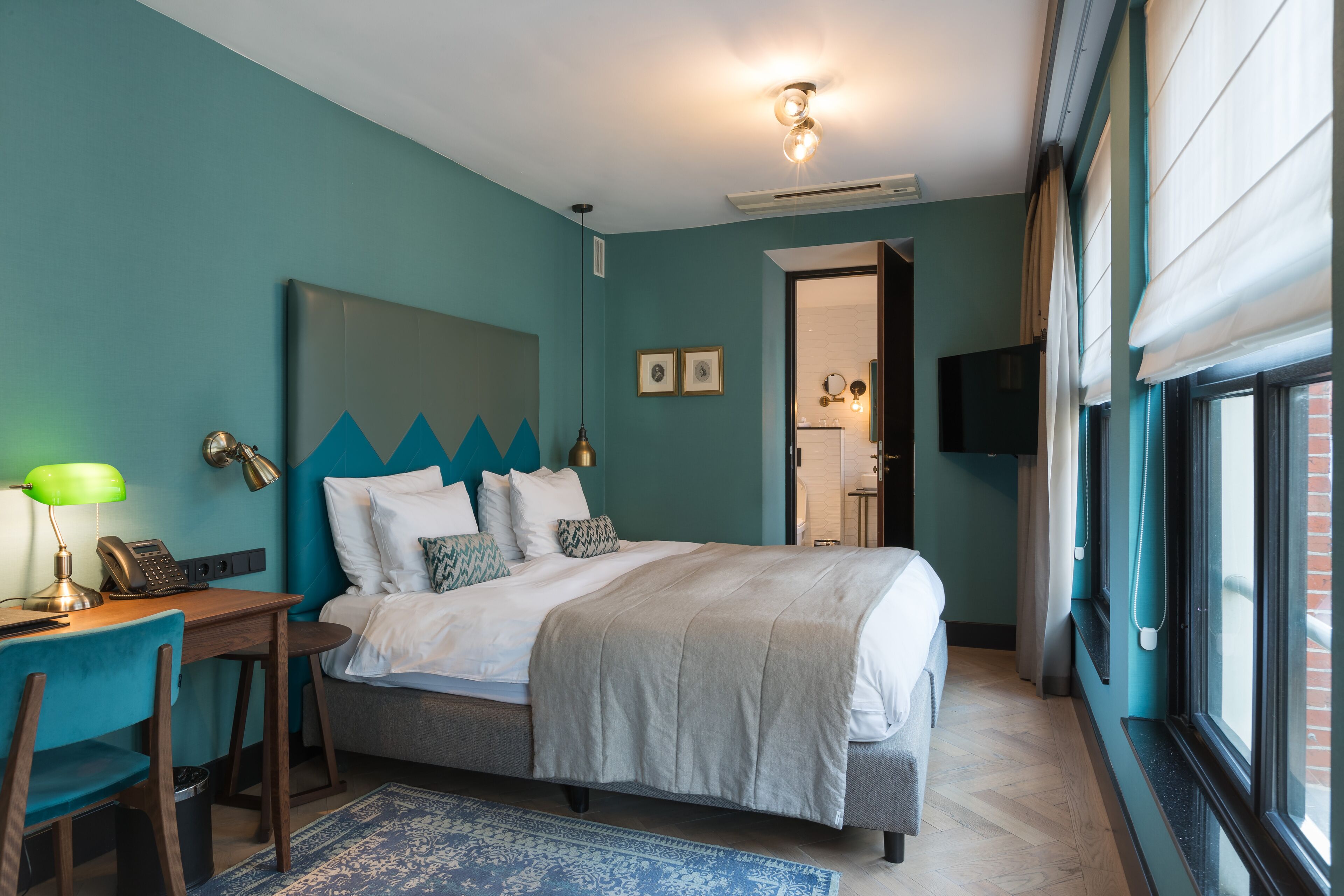 A bedroom with a bed, desk, chair, and a large window with curtains.