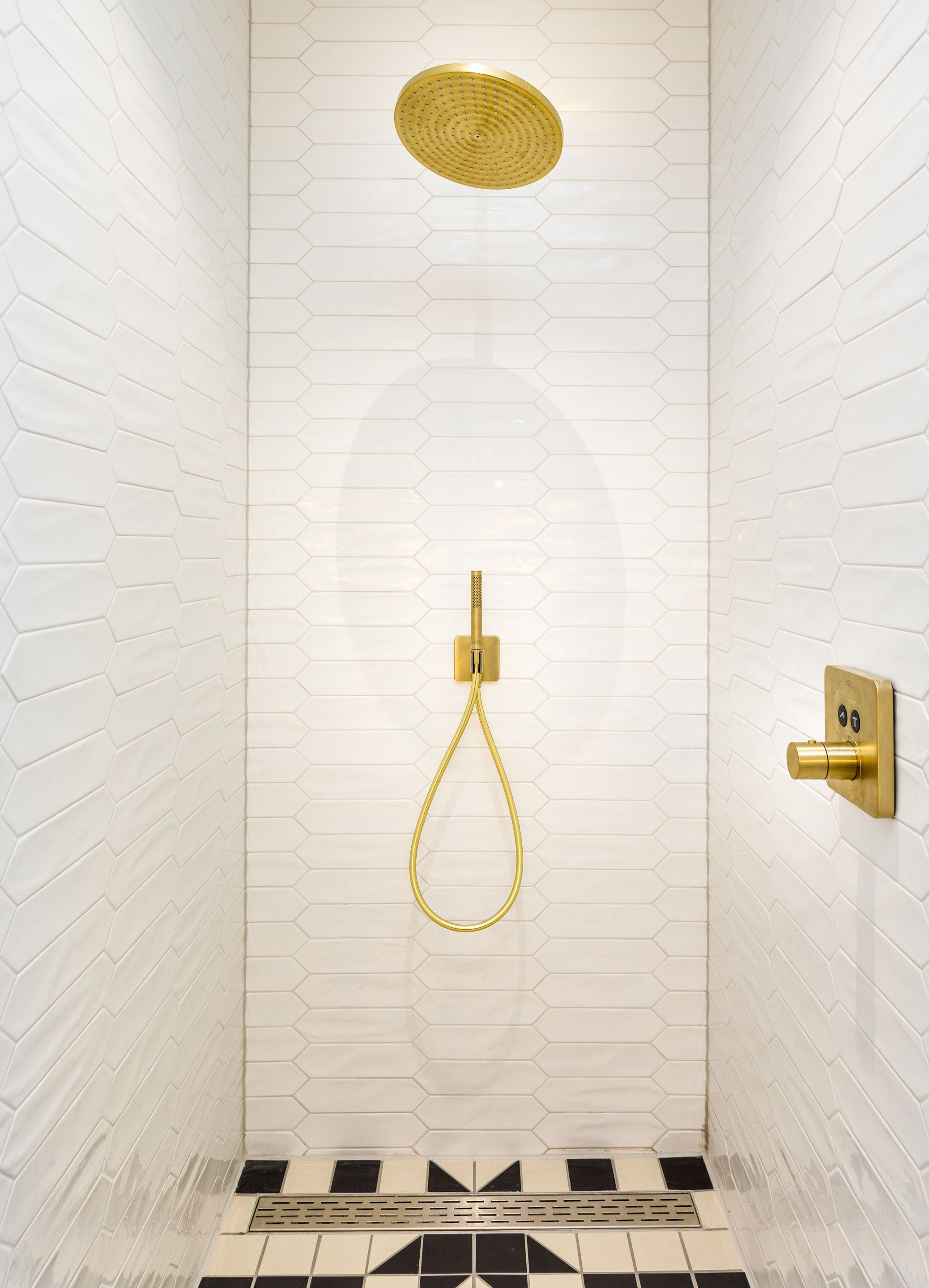 A shower with a gold showerhead and control panel, set against a white hexagonal tile wall.