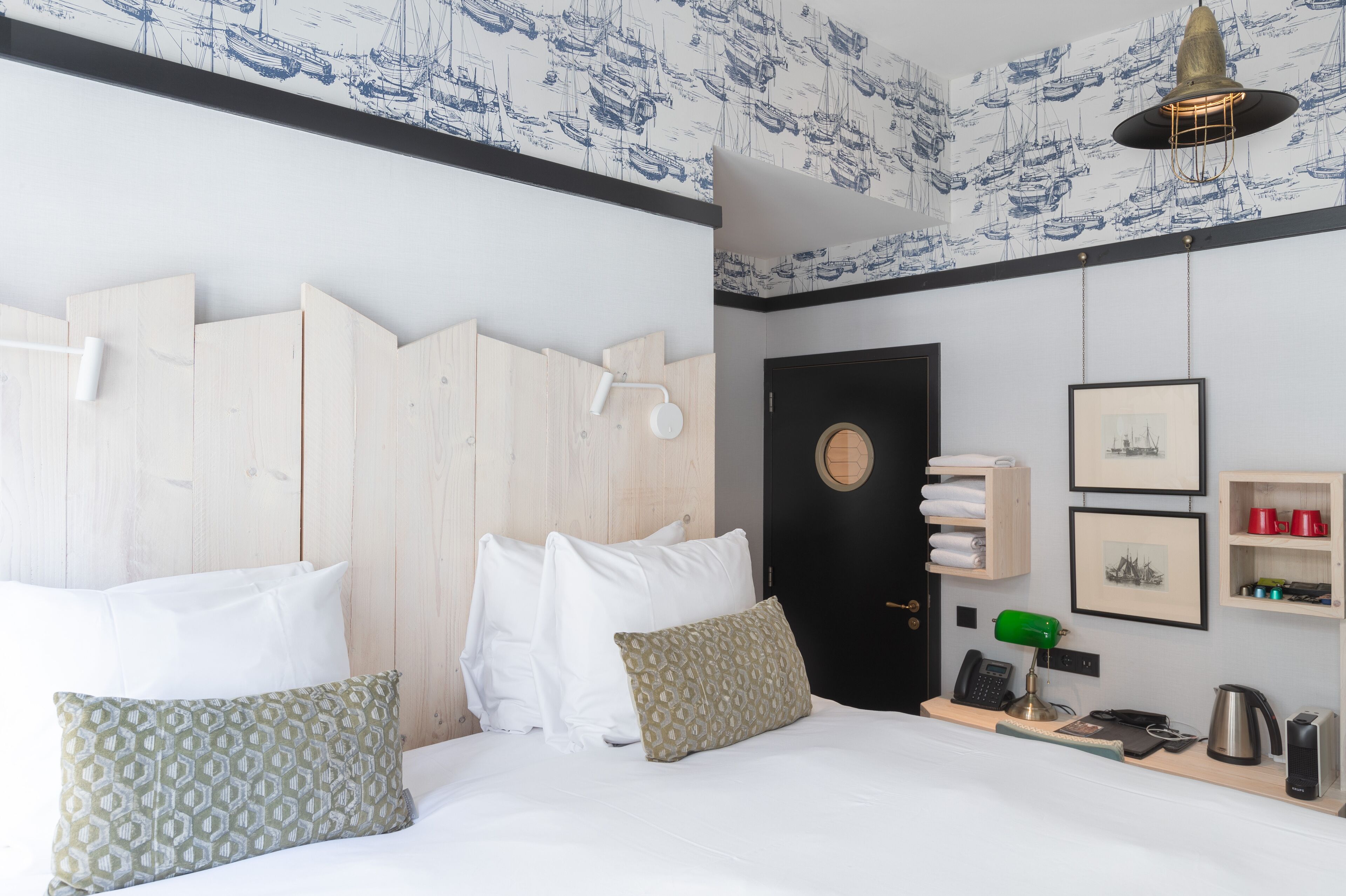 A hotel room with a wooden headboard, white bedding, a desk with a lamp, a phone, a kettle, and wall-mounted shelves with towels and framed pictures.