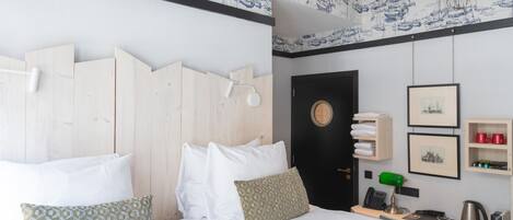 A hotel room with a wooden headboard, white bedding, a desk with a lamp, a phone, a kettle, and wall-mounted shelves with towels and framed pictures.
