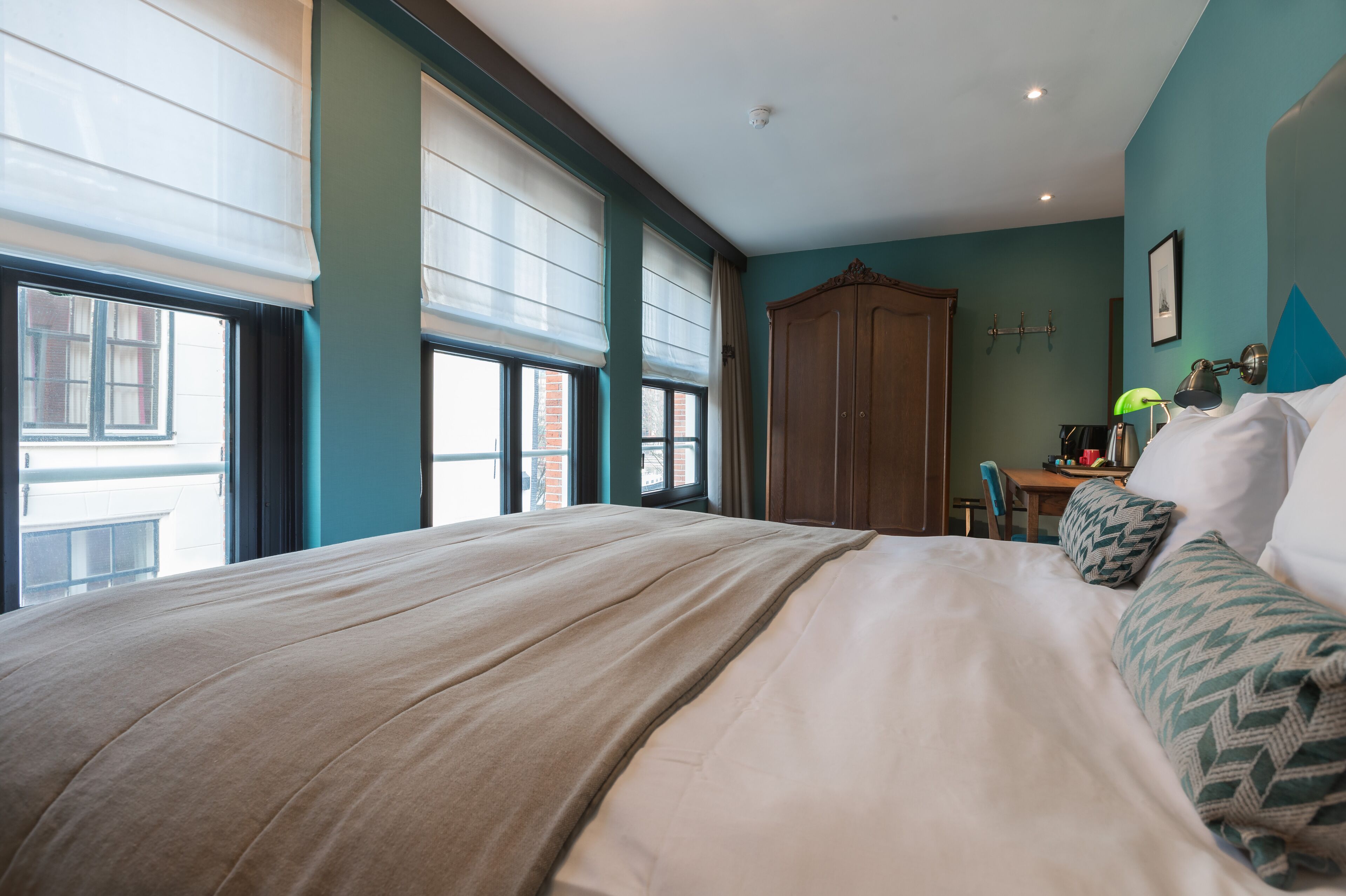 A bedroom with a large bed, a wooden wardrobe, a desk with a chair, and a window with blinds.