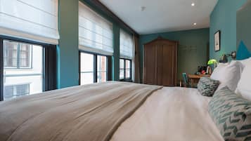 A bedroom with a large bed, a wooden wardrobe, a desk with a chair, and a window with blinds.