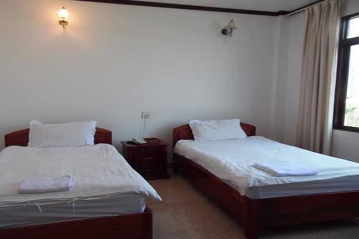 Une chambre avec deux lits simples, chacun doté d’une tête de lit en bois et de draps blancs. Il y a une table de chevet avec une lampe sur chaque lit, et une fenêtre avec des rideaux sur le côté droit.