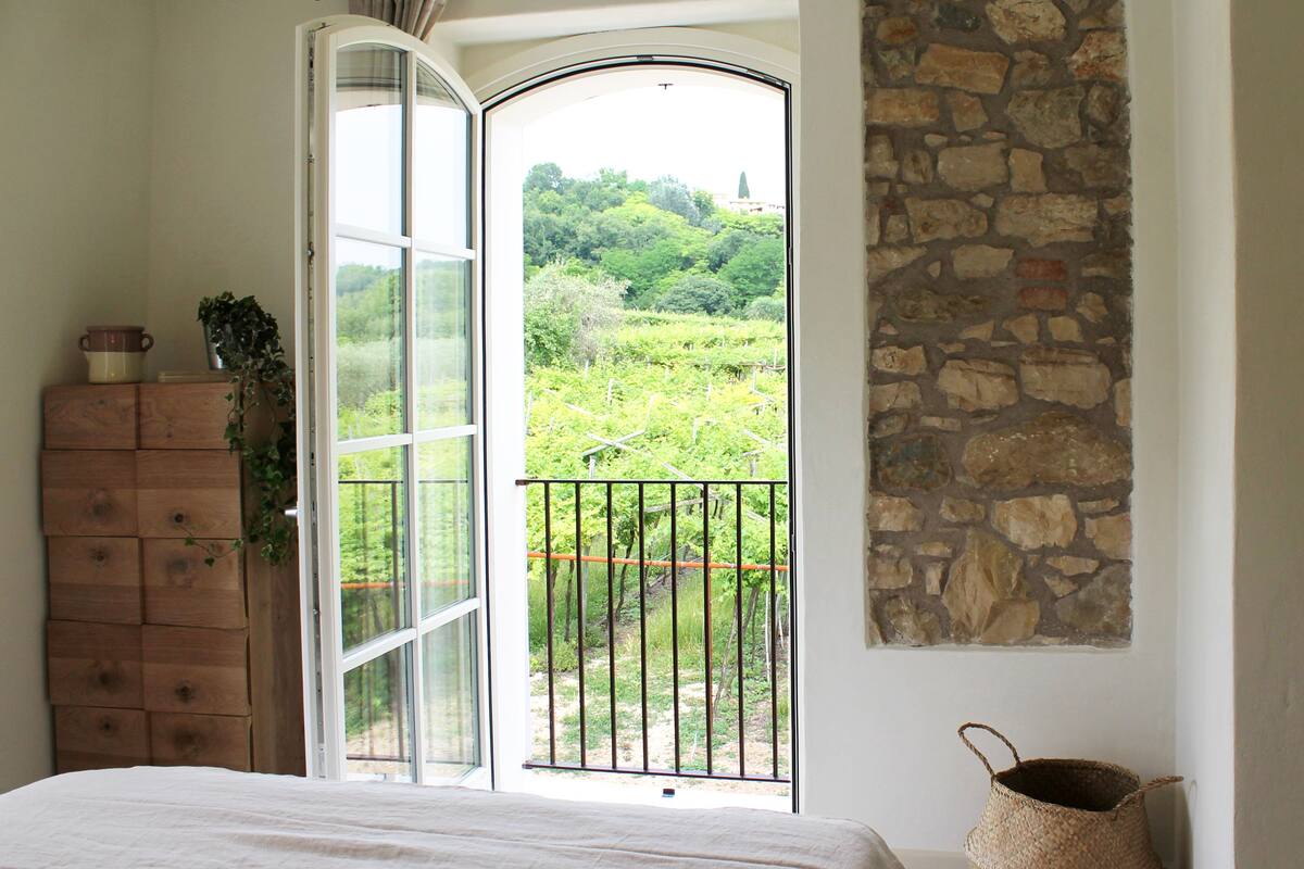 A bedroom with a large window, a wooden dresser, a stone wall, and a woven basket.