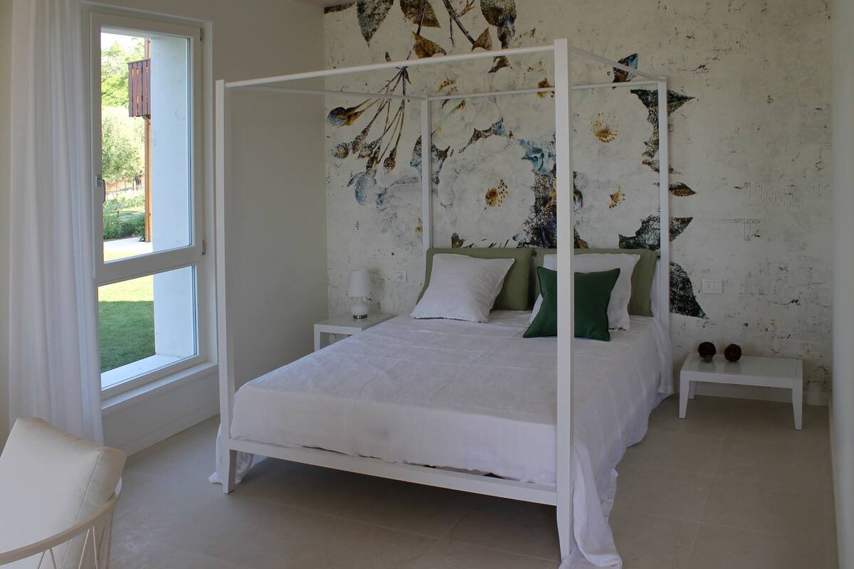 A four-poster bed with white bedding, two bedside tables, and a view of greenery outside.
