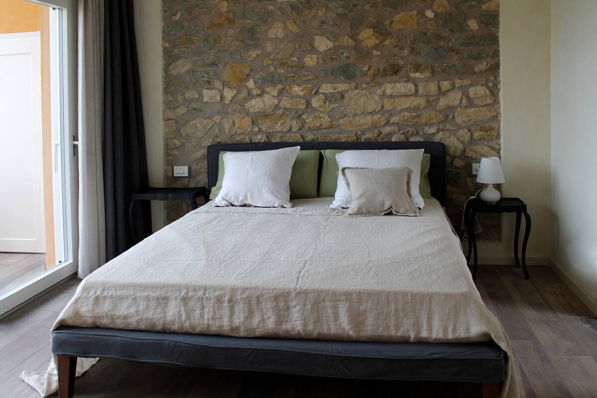 A bed with a stone wall behind it, a window with curtains, and a nightstand with a lamp.