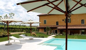 A pool area with lounge chairs and umbrellas, a large house with a tiled roof, and a vineyard in the background. - Ca' Muretta Relais (Bardolino)