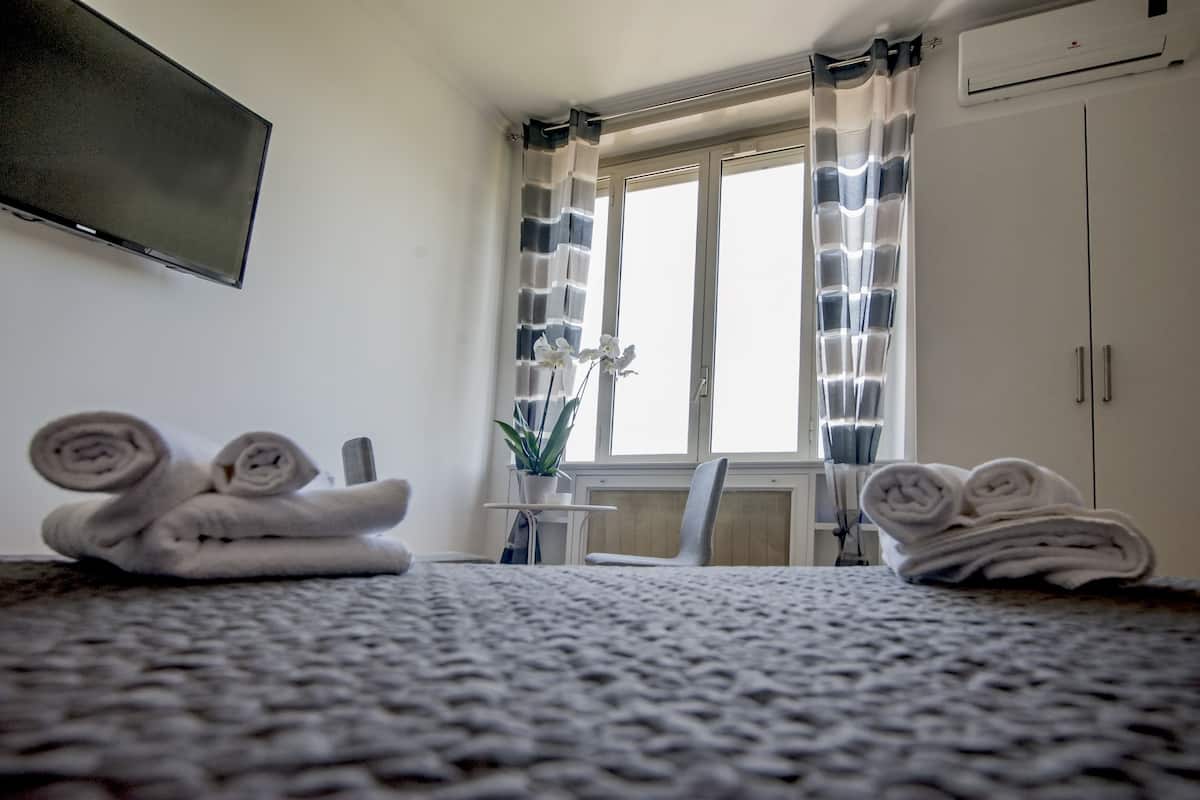 Une chambre d’hôtel avec un lit, un téléviseur fixé au mur, une fenêtre avec des rideaux, une plante en pot et un porte-serviettes.