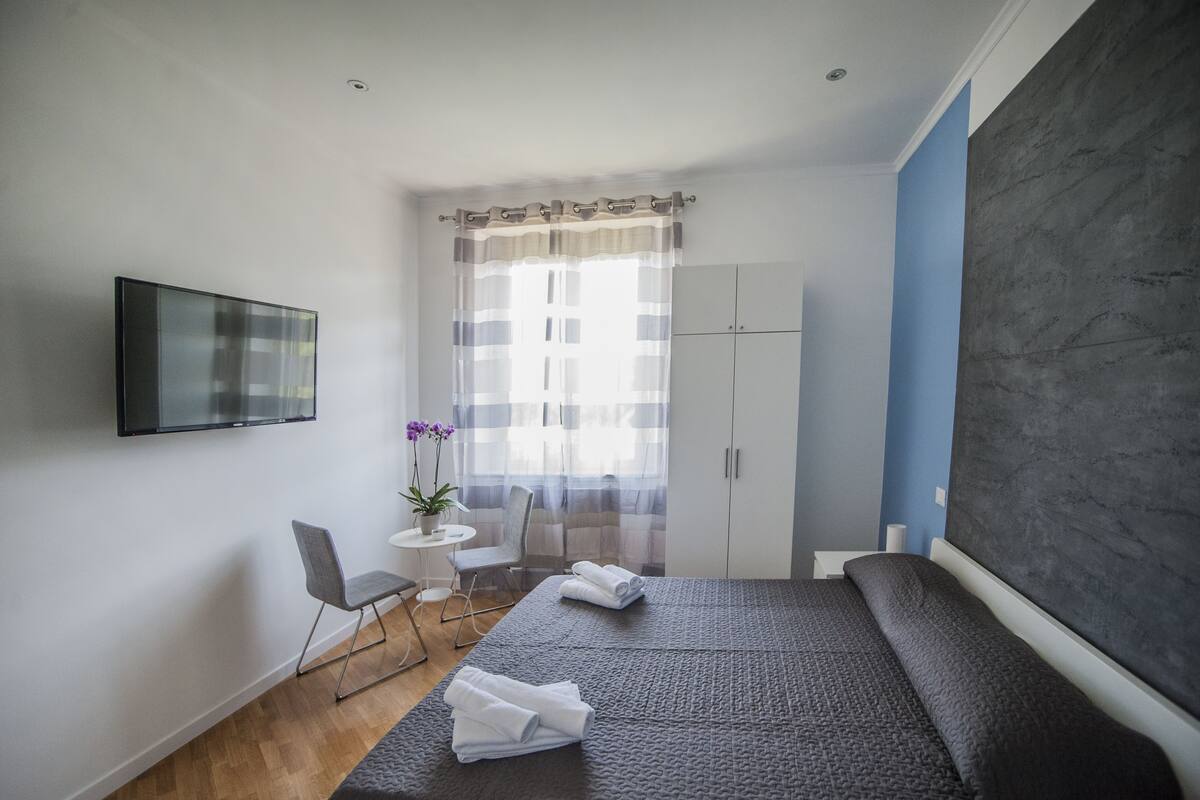 Une chambre d’hôtel moderne, équipée d’un lit, d’un téléviseur fixé au mur, d’une petite table avec une chaise et d’une plante en pot.
