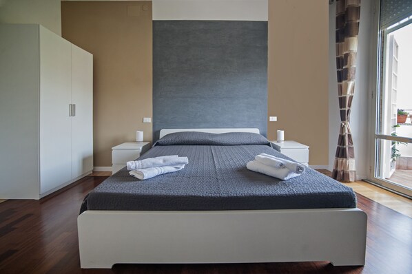 A modern bedroom with a large bed, white nightstands, a wardrobe, and a view of the outdoors through a window. - Guest House Vignola (Rome)