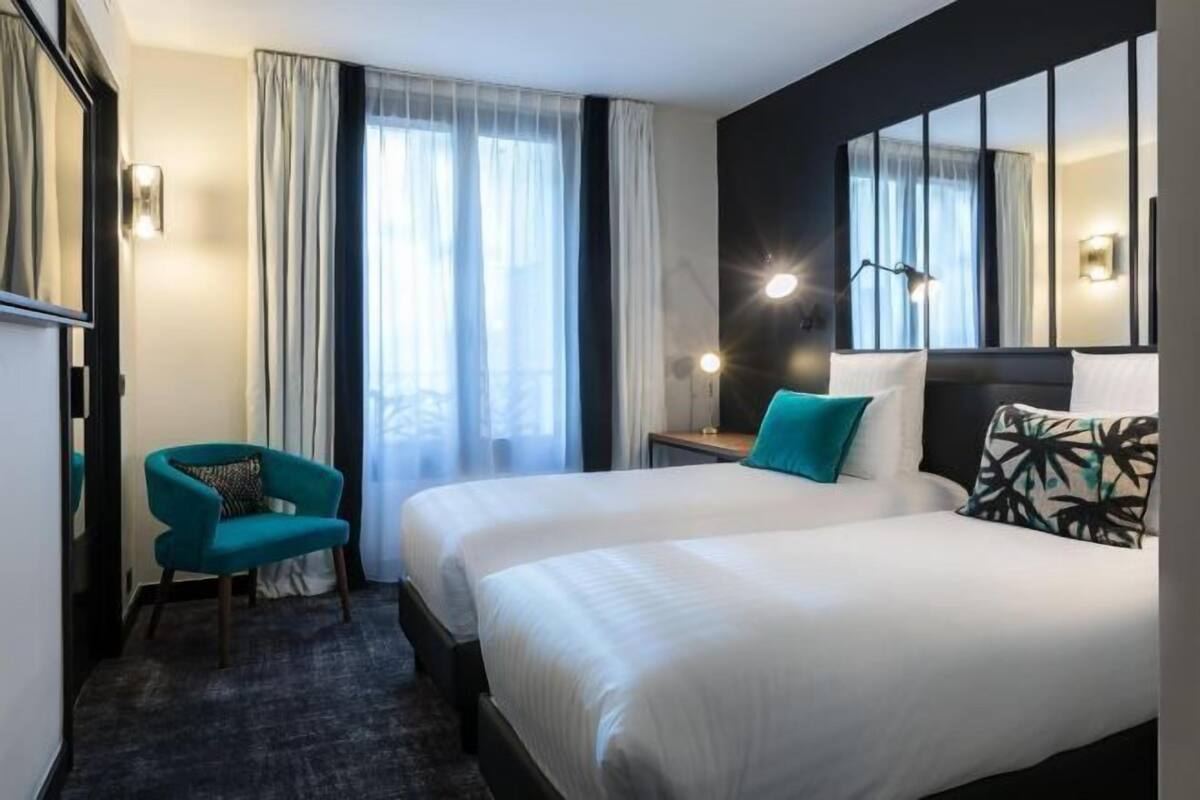 Habitación de hotel con dos camas, un escritorio, una silla y un amplio ventanal con cortinas.