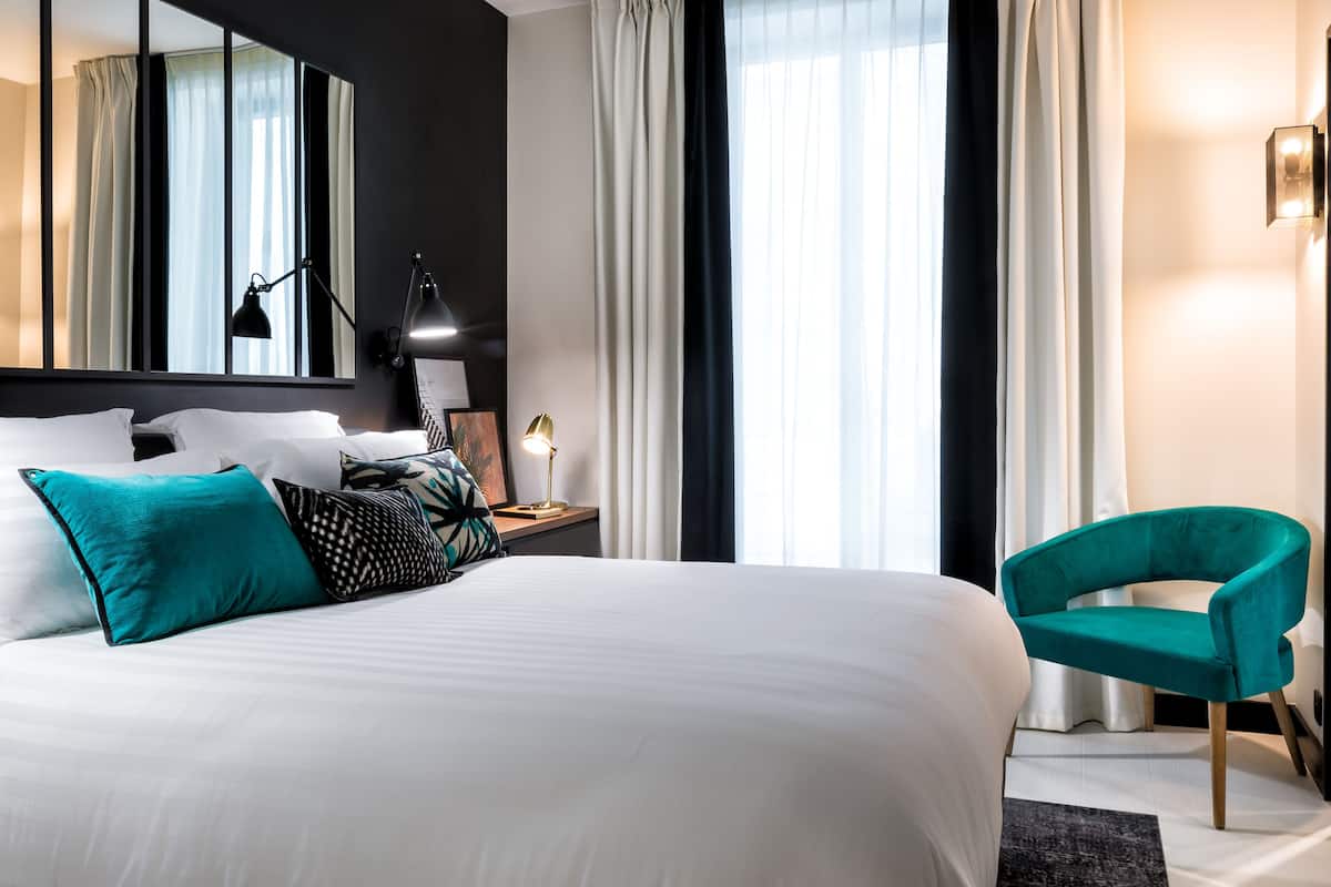 Una habitación de hotel moderna con una cama grande, un sillón verde azulado, una mesita de noche con lámpara y un espejo amplio.