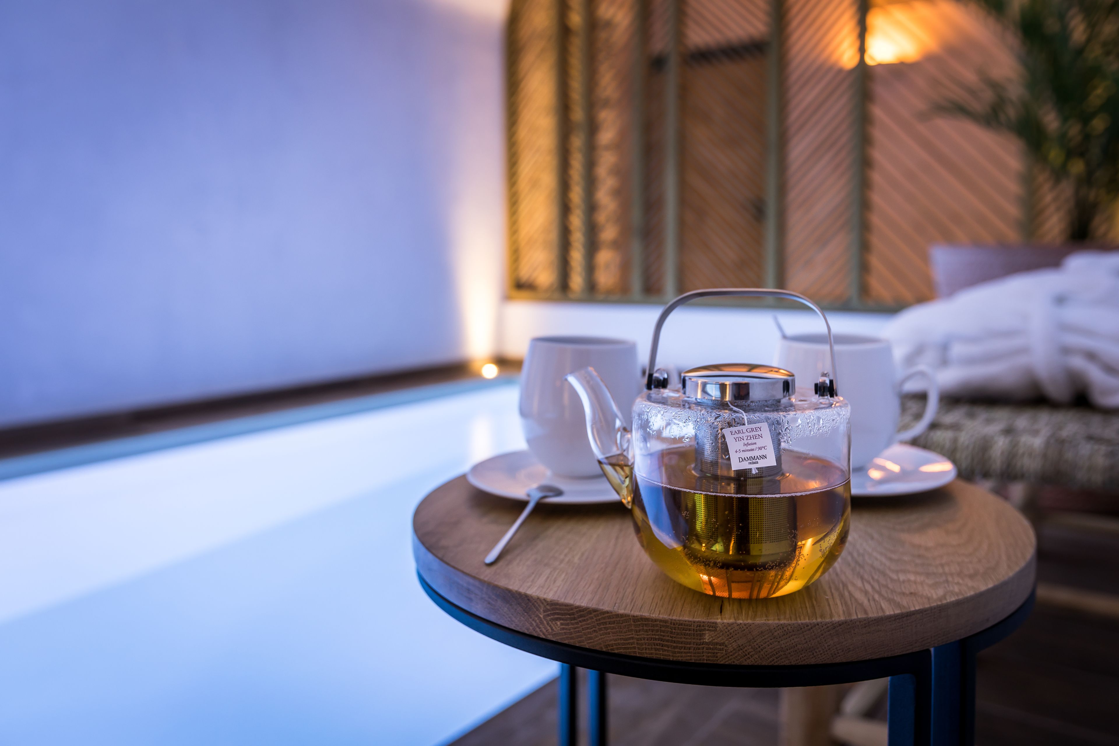 A clear glass teapot with tea on a wooden table, accompanied by a white cup and saucer, in a room with a wicker wall and a bed in the background.