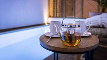 A clear glass teapot with tea on a wooden table, accompanied by a white cup and saucer, in a room with a wicker wall and a bed in the background.