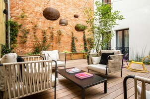 Un patio con mobiliario de mimbre, una mesa con libros, lámparas colgantes de mimbre y una pared de ladrillo cubierta de plantas trepadoras.