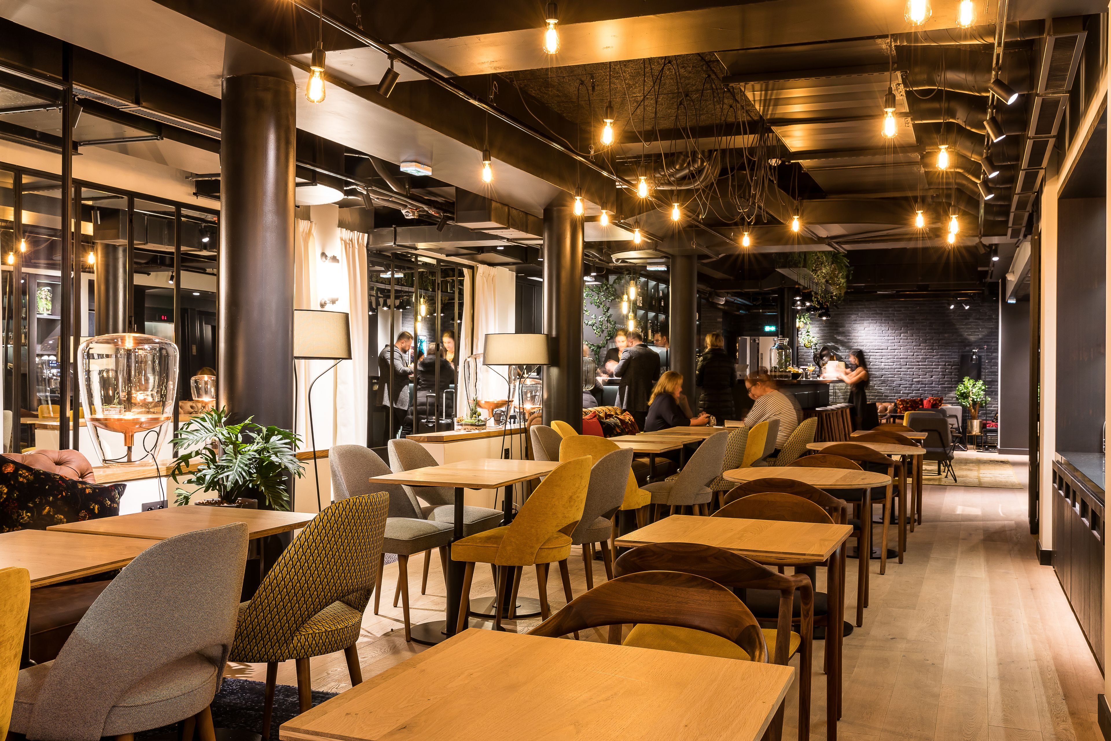 Moderni ravintola, jossa on puupöydät ja -tuolit, esillä olevat kattoputket ja riippuvalaisimet.