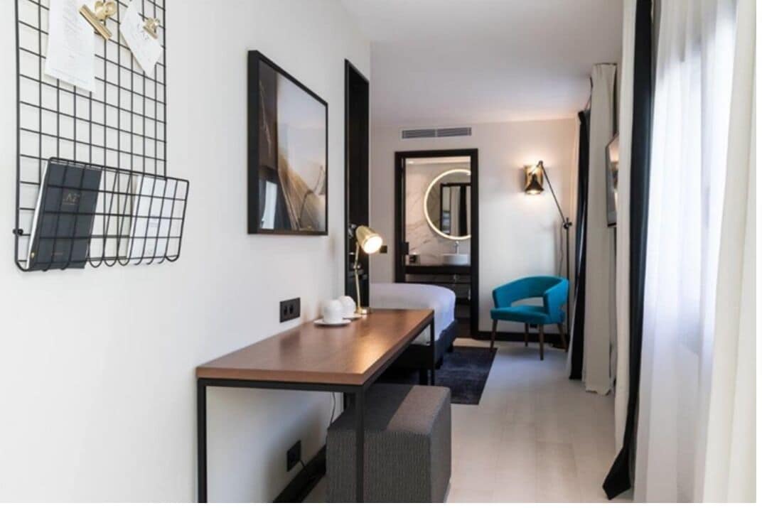 A modern hotel room with a wooden desk, a blue chair, a bed, a mirror, and a wall-mounted light fixture.