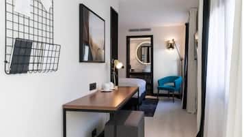A modern hotel room with a wooden desk, a blue chair, a bed, a mirror, and a wall-mounted light fixture.