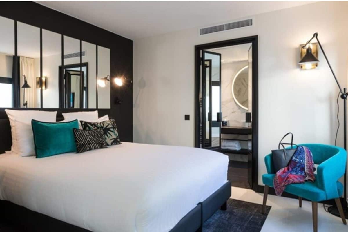 Una habitación de hotel moderna con una cama grande, un sillón azul, un espejo y una lámpara de pared.