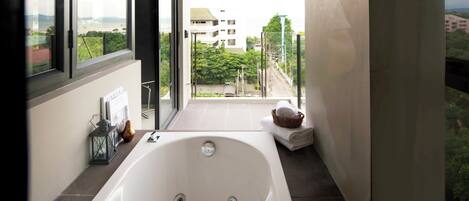 A modern bathroom with a large bathtub, a window with a city view, and a small table with towels and a lantern.