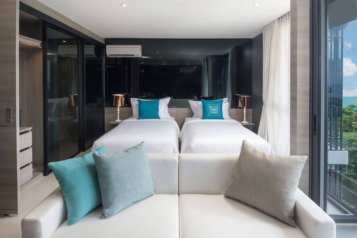 A modern hotel room with two beds, a sofa, and a large window offering a view of the city.