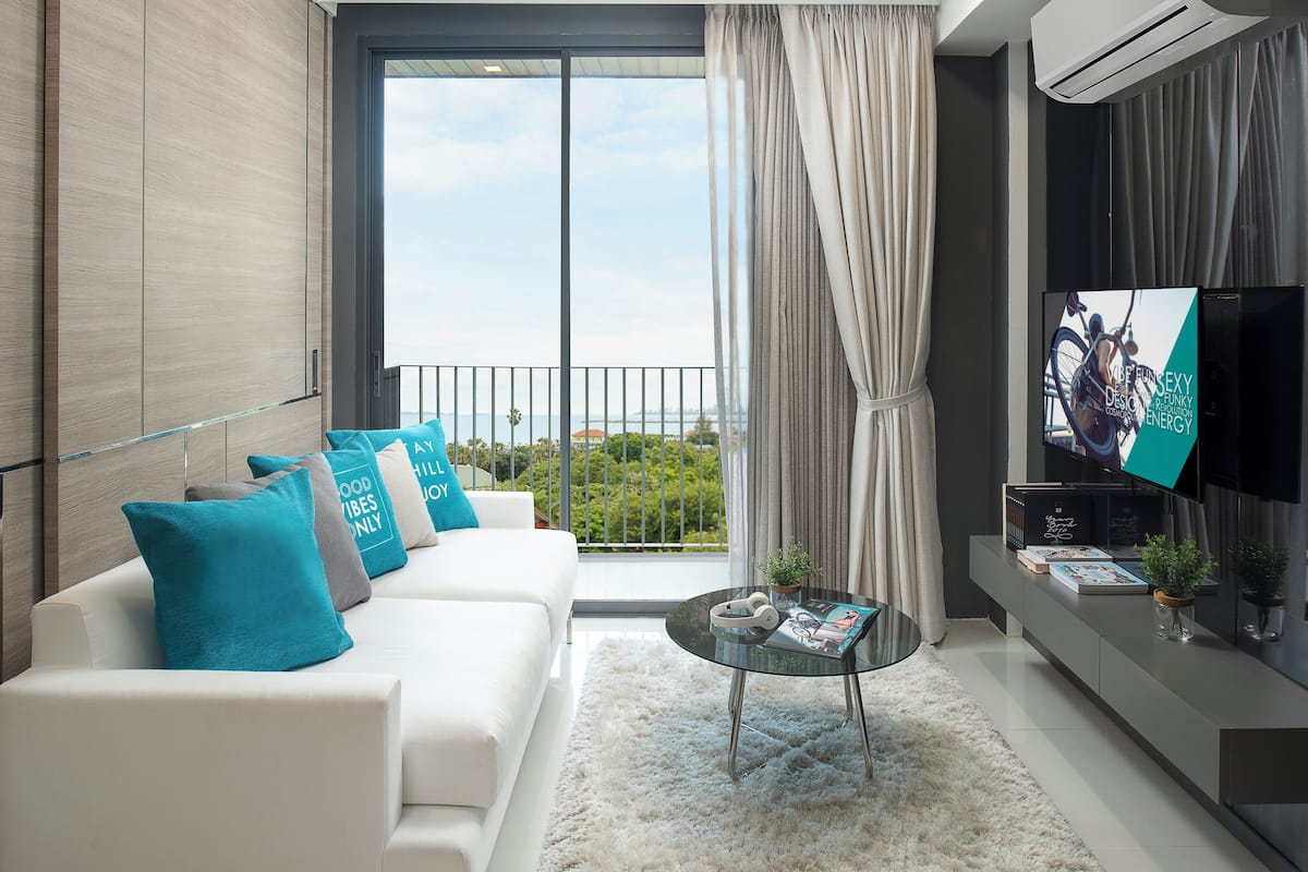 A modern living room with a white sofa, teal cushions, a glass coffee table, and a flat-screen TV on a black stand.